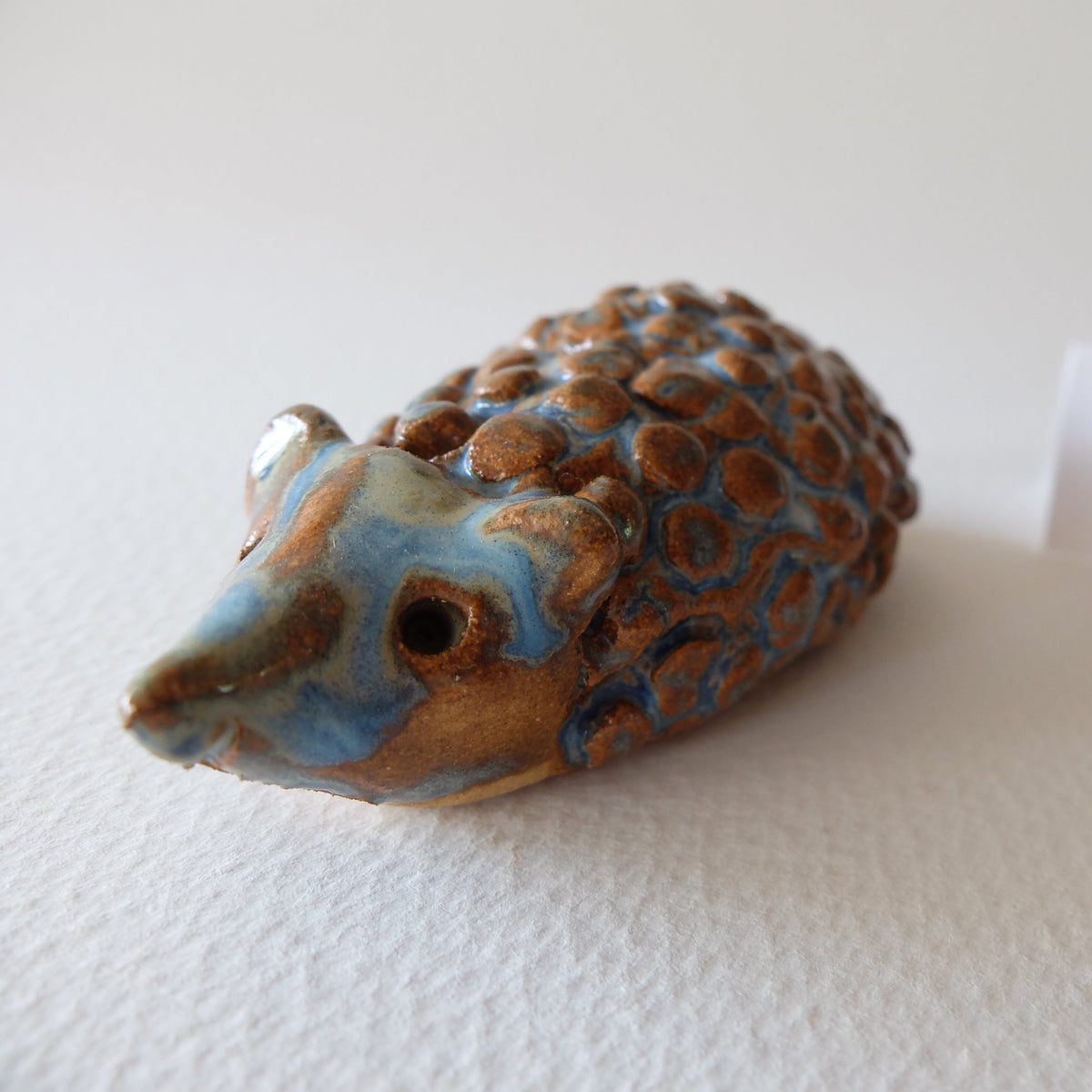 Handcrafted ceramic hedgehog figurine "The Bold Blue-Raspberry Hedgehog (#18)" in stoneware by Nikolaj Jesper Cyon. Features a confident posture with rounded bumpy quills in brown glaze and vibrant cobalt blue glaze around the face, ears, and back.