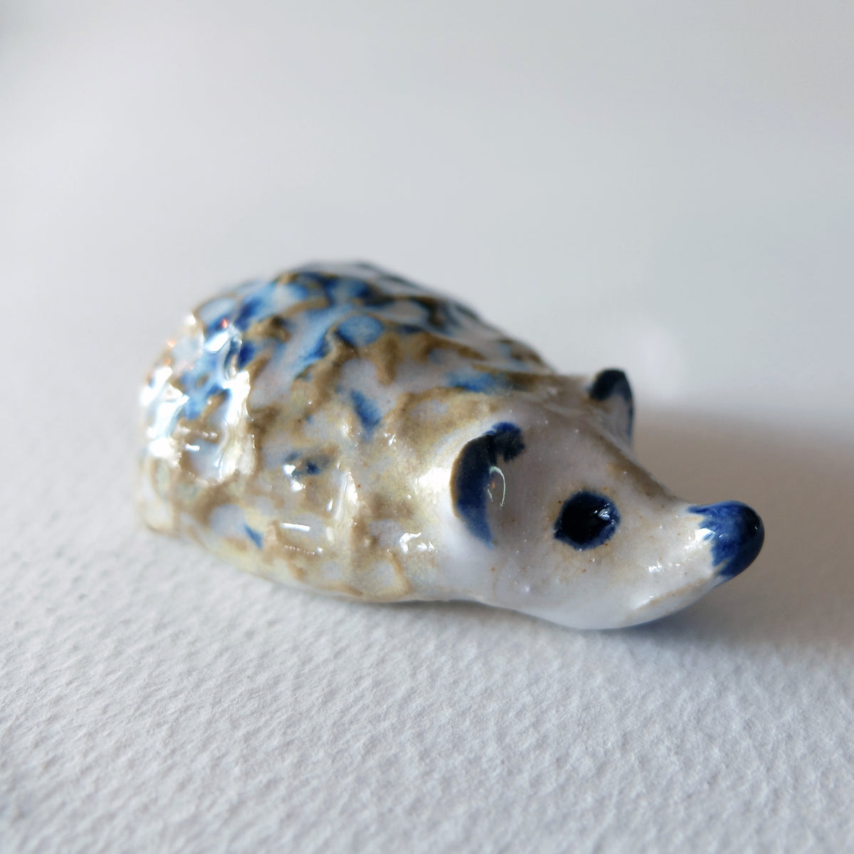 Handcrafted ceramic hedgehog figurine "The Frosty Little Hedgehog (#24)" in stoneware by Nikolaj Jesper Cyon. Tiny, rugged and buckled hedgehog sculpture with a textured pattern of deep blue and warm ochre on a snowy white glaze.