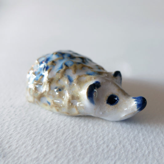 Handcrafted ceramic hedgehog figurine "The Frosty Little Hedgehog (#24)" in stoneware by Nikolaj Jesper Cyon. Tiny, rugged and buckled hedgehog sculpture with a textured pattern of deep blue and warm ochre on a snowy white glaze.