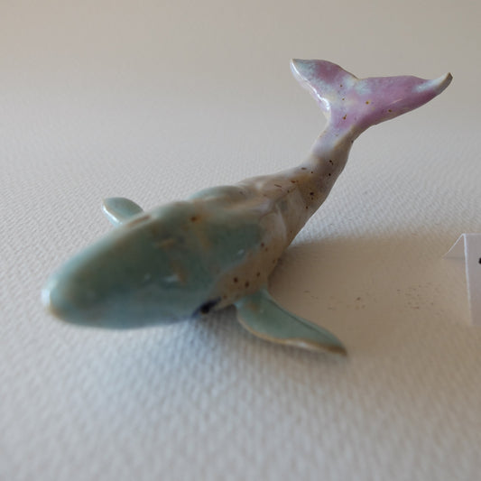 Handcrafted ceramic whale figurine "The Pastel Whale (#10)" in stoneware by Nikolaj Jesper Cyon. Glazed in soft pastel tones with a light blue-green body, speckled beige back, and a raised lavender-pink tail fin.