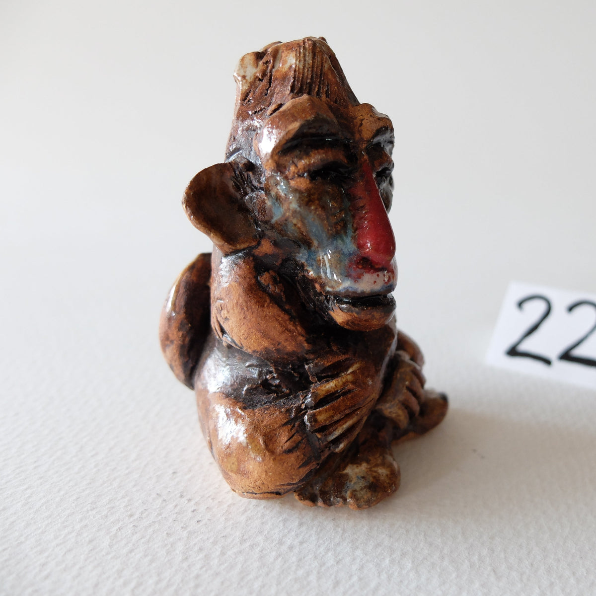 Handcrafted ceramic mandrill figurine "The Pensive Mandrill (#22)" in stoneware by Nikolaj Jesper Cyon. Seated monkey sculpture in warm brown glazes featuring a distinct red nose and detailed fur texture.