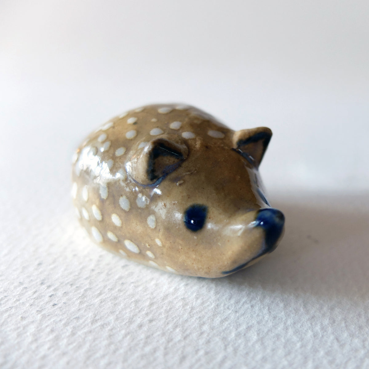 Handcrafted ceramic hedgehog figurine "The Polka Dotted Hedgehog (#25)" in stoneware by Nikolaj Jesper Cyon. Small rounded hedgehog sculpture in a warm tan glaze with delicate white polka dots and cobalt blue facial features.