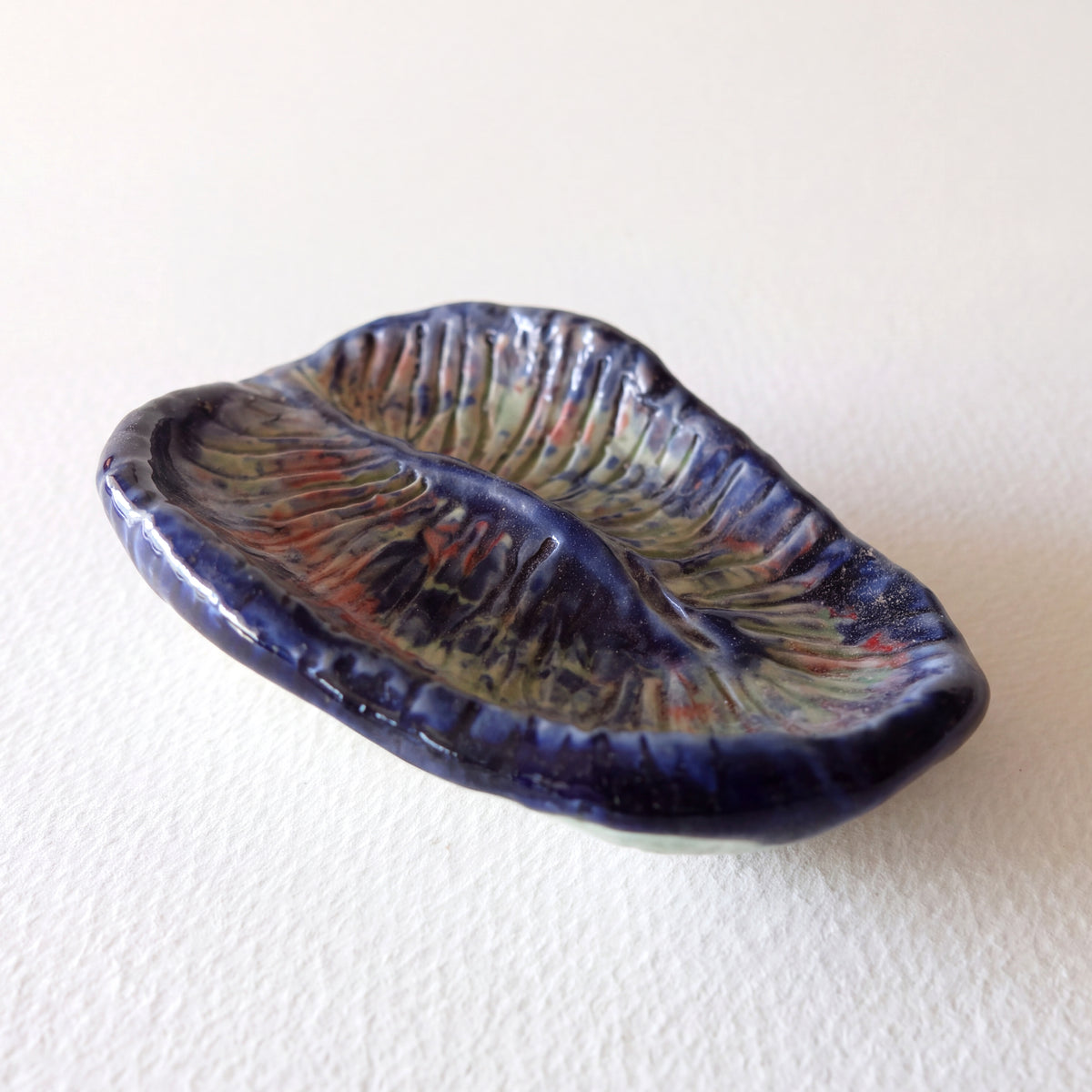 Handcrafted ceramic art object "The Pollen Grain Vessel (#19)" in stoneware by Nikolaj Jesper Cyon. Oval concave dish inspired by microscopic pollen grains, with carved radial grooves glazed in deep cobalt blue, pale green, and coral red,