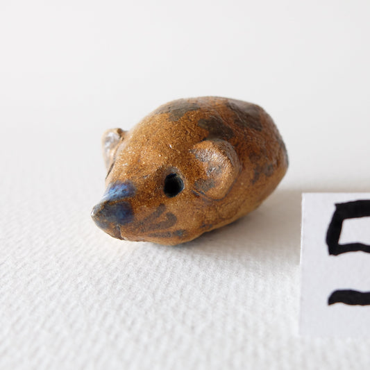 Handcrafted ceramic hedgehog figurine "The Chestnut Hedgehog (#5)" in stoneware by Nikolaj Jesper Cyon. Features a warm, speckled brown and ochre body with dark decorative spots and a touch of blue glaze on the pointed snout, captured in a three-quarter front view.