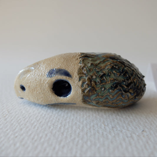 Handcrafted ceramic chimeric figurine "The Hedgehog Bird (#9)" in stoneware by Nikolaj Jesper Cyon. Features a smooth, light beige bird-like head with dark blue eyes and beak, combined with a textured, quilled hedgehog-like back in green, brown, and blue glazes.